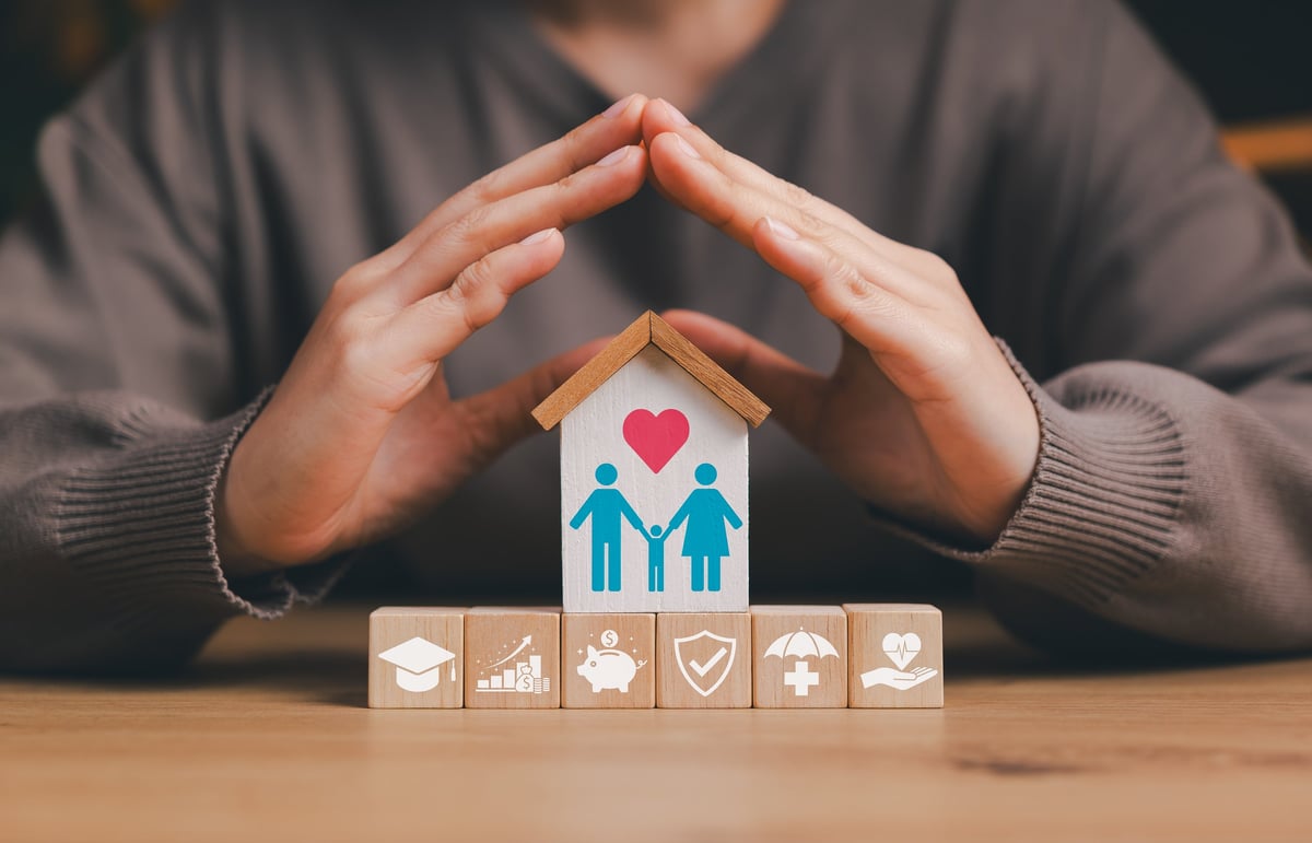 Hands protecting a wooden house model with family figures and a heart symbol, representing family financial protection
